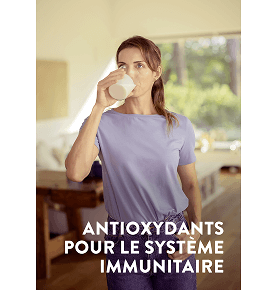 Une femme boit un verre blanc de Pediasure vanille dans son salon avec les mots avec des antioxydants pour soutenir votre système immunitaire