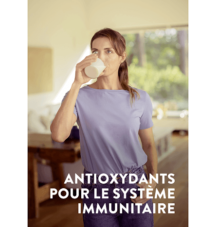 Une femme boit un verre blanc de Pediasure vanille dans son salon avec les mots avec des antioxydants pour soutenir votre système immunitaire