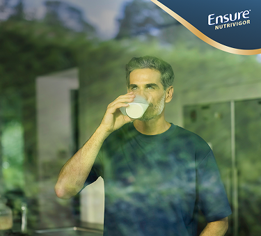 Een man drinkt een voedingssupplement terwijl hij uit het raam naar een grasveld kijkt