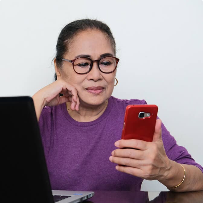 Elderly woman wearing glasses looking at the phone in hand with a laptop on the side