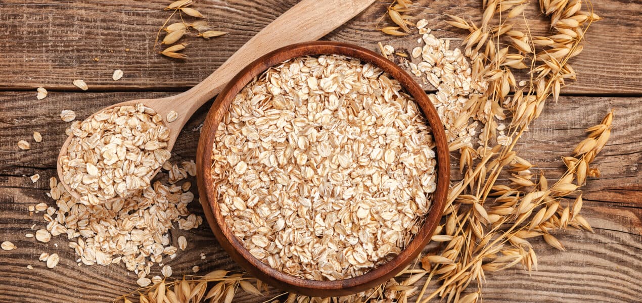 A wooden bowl and spoon full of whole grain oats with dried wheats on the sides