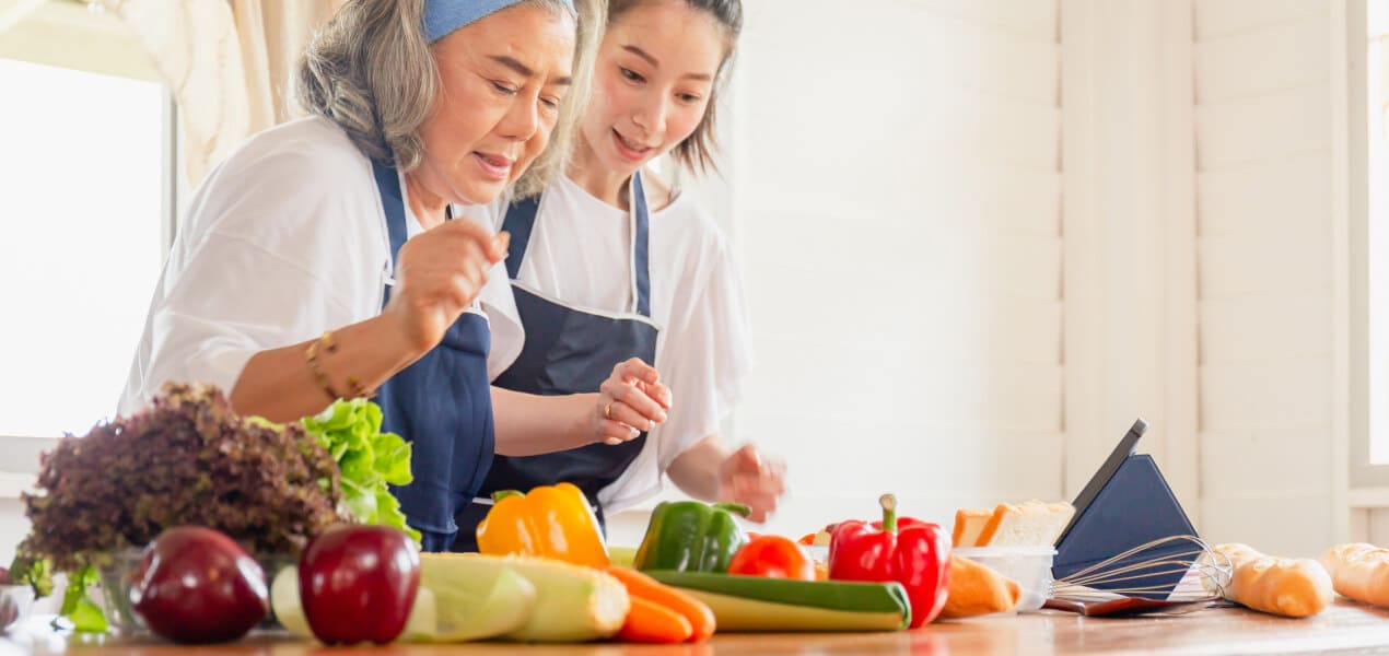 An elderly and young woman wearing aprons with different healthy ingredients in front of them